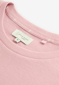 Vaaleanpunainen, teksturoitu neulepaita, jossa on pyöreä kaula-aukko. Sisäkauluksessa on pieni etiketti, jossa lukee "By Ted Baker" ja kokolappu.