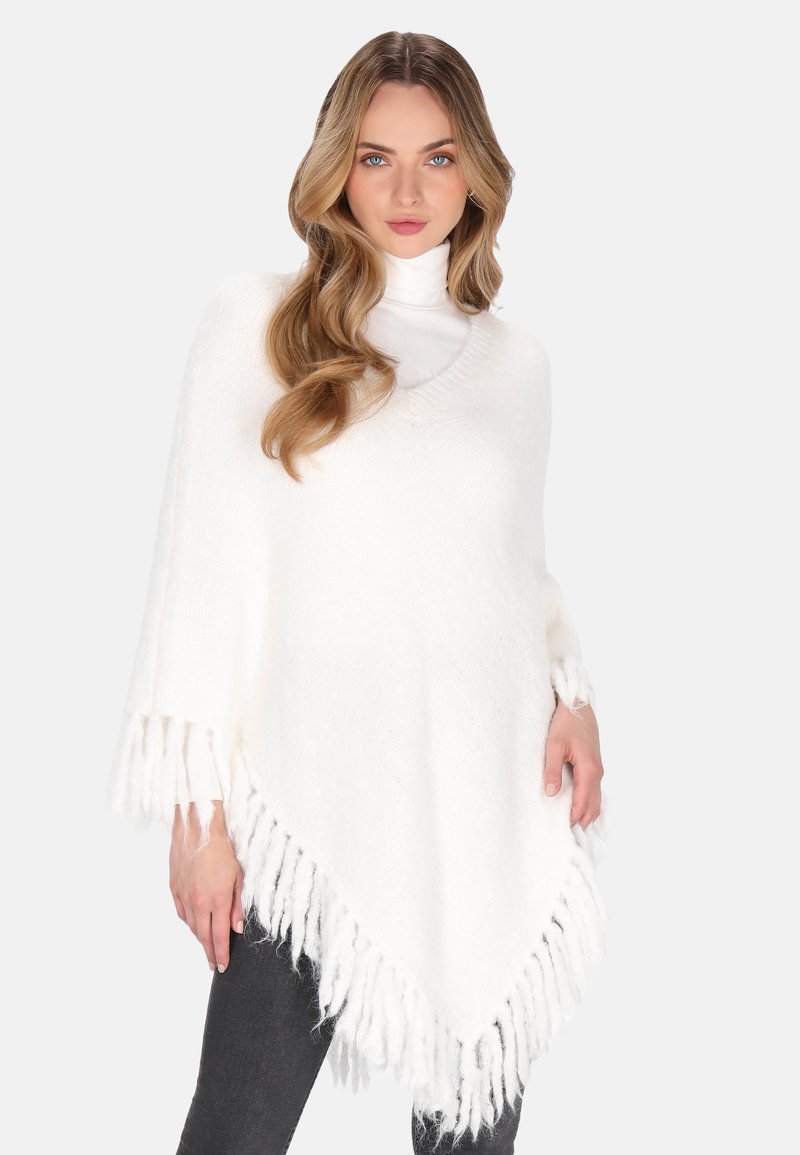 Donna con lunghi capelli ondulati che indossa un poncho bianco con frange sopra un dolcevita bianco e pantaloni scuri, in piedi davanti a uno sfondo neutro.
