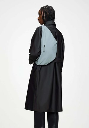 Persona con capelli intrecciati indossa un lungo cappotto nero e uno zaino a tracolla grigio chiaro, di profilo su sfondo neutro.