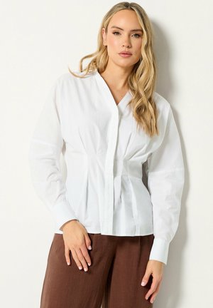 Vrouw met lang blond haar, die een witte blouse met knopen en een bruine broek draagt, staand tegen een effen lichte achtergrond.
