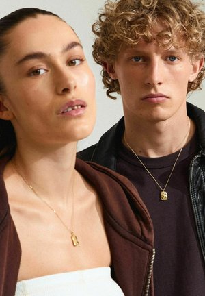 Zwei Models stehen der Kamera gegenüber und tragen goldene Halsketten. Die Frau hat lange Haare und trägt eine braune Jacke über einem weißen Oberteil. Der Mann trägt ein dunkles Hemd.