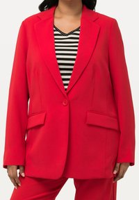 Femme portant un blazer rouge vif à un seul bouton sur un chemisier à col en V rayé noir et blanc, et un pantalon rouge assorti.