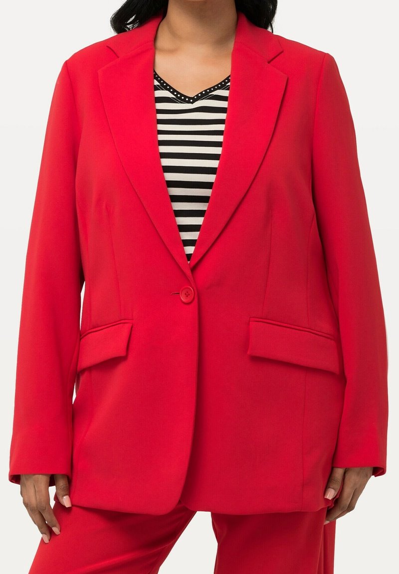 Femme portant un blazer rouge vif à un seul bouton sur un chemisier à col en V rayé noir et blanc, et un pantalon rouge assorti.