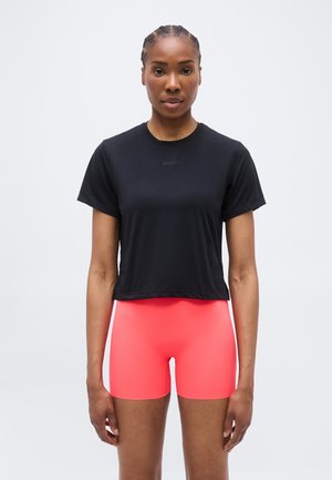 Žena u crnoj kratkotrajnoj majici i svijetlo koraljno ružičastim uskim kratkim hlačama stoji uspravno s neutralnim izrazom lica na jednobojnoj pozadini.