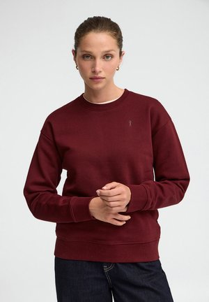 Donna dalla pelle chiara e occhi verdi che indossa una felpa color bordeaux e jeans scuri, in piedi davanti a uno sfondo semplice.