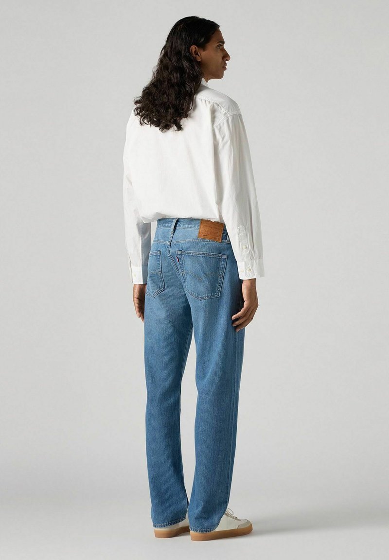 Jeans en denim délavé avec une coupe droite, dotés d'une patch en cuir marron au niveau de la taille et d'un léger dégradé. Associés avec une chemise blanche à boutons.