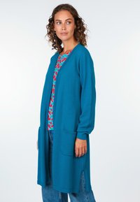 Teal strukturierte Strickjacke mit langen Ärmeln, offener Front und zwei Vordertaschen. Kombiniert mit einem blumenmusternden Oberteil und blauen Jeans.