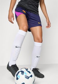 Shorts Nike Barcelona en bleu marine avec des accents violets, associés à de hautes chaussettes blanches et des chaussures de football noires. Un ballon de football texturé est à proximité.