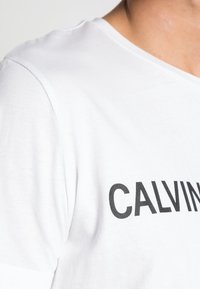Bílý bavlněný tričko s kulatým výstřihem, krátkými rukávy a výrazným černým textem "CALVIN" na přední straně. Hladký materiál, volný střih.