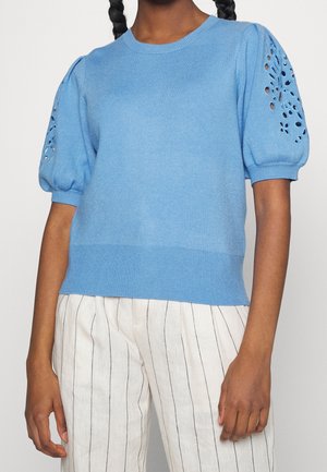 Mujer con suéter azul claro de manga corta con detalle floral recortado en las mangas y pantalones blancos con finas rayas negras verticales.
