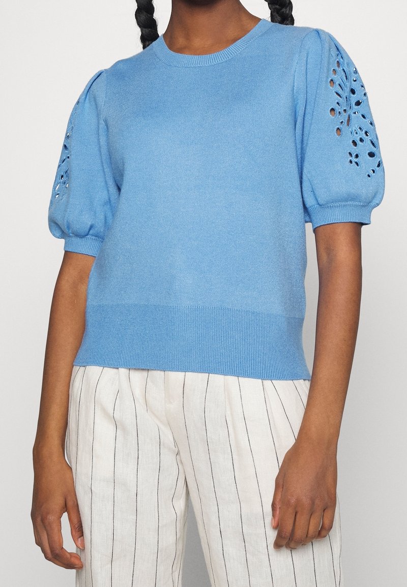 Femme portant un pull à manches courtes bleu clair avec des détails en découpe florale sur les manches et un pantalon blanc avec de fines rayures noires verticales.