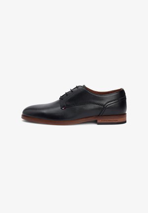 Black leather dress shoe with smooth texture, rounded toe, and six-eyelet lace-up design. Features a brown heel with logo detail.