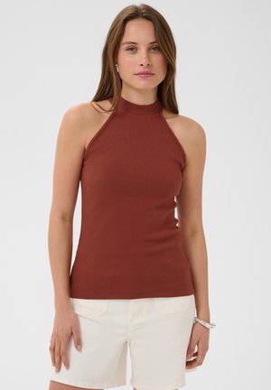 Femme portant un haut côtelé sans manches couleur rouille et un short blanc, debout avec une expression neutre devant un fond uni.