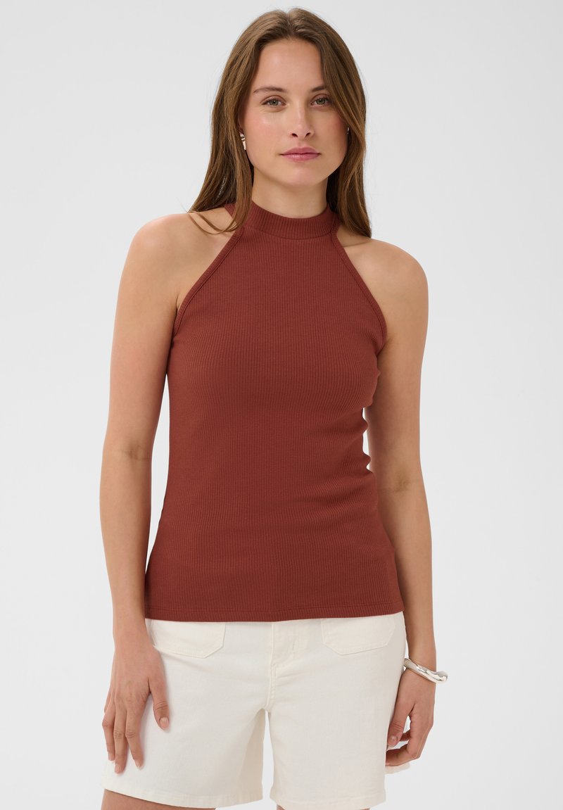 Femme portant un haut côtelé sans manches couleur rouille et un short blanc, debout avec une expression neutre devant un fond uni.