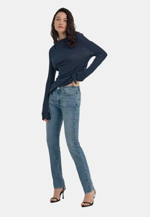 Maglia a maniche lunghe blu navy con dettaglio laterale annodato, abbinata a jeans slim-fit azzurri e sandali neri con tacco alto. Texture del tessuto lisce.