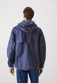 Lehká námořnická bunda s kapucí, vyrobená z texturované látky a se stahovacími manžetami, určená pro volný čas. Zadní pohled ukazuje detaily švů.