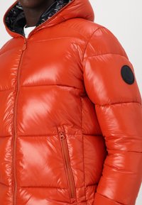 Red puffer jacket with glossy finish, zip closure, and an external pocket. Features a black logo patch on the left upper arm.