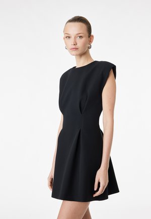 Jeune femme aux cheveux tirés en arrière portant une robe noire sans manches avec des épaules structurées, sur un fond blanc uni.