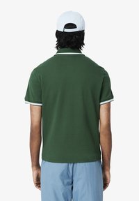 Zielony polo z białymi akcentami na kołnierzyku i rękawach; noszony z jasnoniebieskimi szortami; widziany od tyłu. Materiał bawełniany, casualowe dopasowanie.