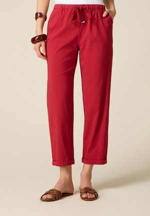 Vrouw met rode broek met koordje en omgeslagen zomen, bruine gevlochten sandalen, een wit topje en flinke bruine armbanden om één pols.