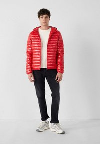 Giacca imbottita rossa con cappuccio, chiusura con zip e una texture lucida, abbinata a una felpa chiara, pantaloni scuri e sneakers beige.