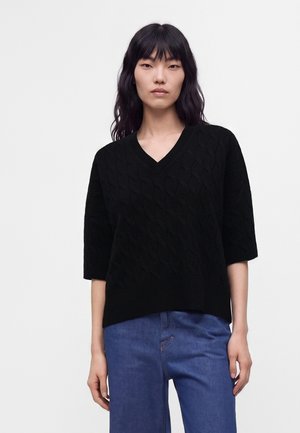 Kvinde med langt sort hår iført en sort kabelstrikket V-hals sweater med albuelange ærmer og blå jeans mod en ensfarvet baggrund.