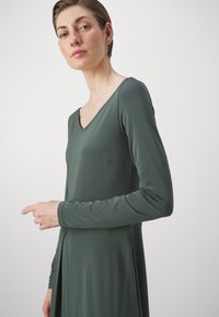 Robe à manches longues de couleur vert foncé, avec un décolleté en V et une texture lisse. La silhouette est ajustée avec un ourlet évasé et des détails sur les manches.
