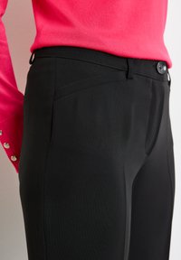 Schwarze, schmal geschnittene Hosen mit glatter Textur, ausgestattet mit seitlichen Taschen und einem Knopfverschluss. Der Bund weist ein dezentes Designelement auf.