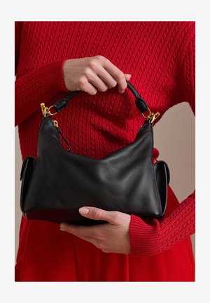 Άτομο με κόκκινο πουλόβερ με πλέξη κρατά μαύρη δερμάτινη τσάντα με χρυσά μεταλλικά στοιχεία από την επάνω λαβή, υποστηρίζοντας τη βάση με το άλλο χέρι.