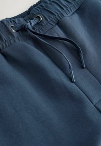 Marineblauwe joggingbroek van zacht materiaal. Elastische tailleband met verstelbare trekkoorden en metalen oogjes. Soepele textuur; geen zichtbare patronen.
