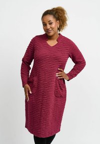 Bordeauxrotes Kleid mit einem strukturierten wellenförmigen Muster, langen Ärmeln, V-Ausschnitt und zwei vorderen Taschen. Glattes Material mit einer weichen Oberfläche.