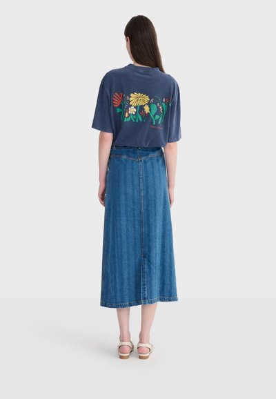 Femme portant un t-shirt bleu marine avec un motif floral coloré au dos, une jupe midi en denim bleu taille haute et des sandales beiges, debout de dos.
