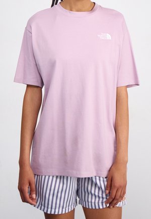 Osoba ubrana w luźną, jasnofioletową koszulkę North Face i białe szorty z ciemnymi pionowymi paskami, stojąca na jednolitym tle.