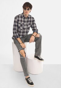 Camicia a quadri con tonalità grigio, blu e rosso; jeans strappati grigi; sneakers nere; occhiali da sole rotondi. Soggetto seduto su sgabelli cilindrici bianchi.