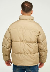 Beige gepolsterte Jacke mit hohem Kragen, ausgestattet mit gesteppten horizontalen Paneelen und elastischen Bündchen. Über blauen Jeans getragen.
