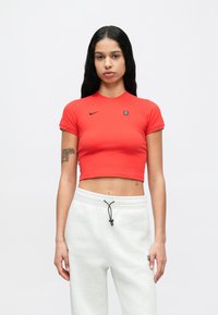 Červené krátke tričko s okrúhlym výstrihom a krátkymi rukávmi, s čiernym logom Nike. Nosené so sivými teplákmi, bežná textúra látky.