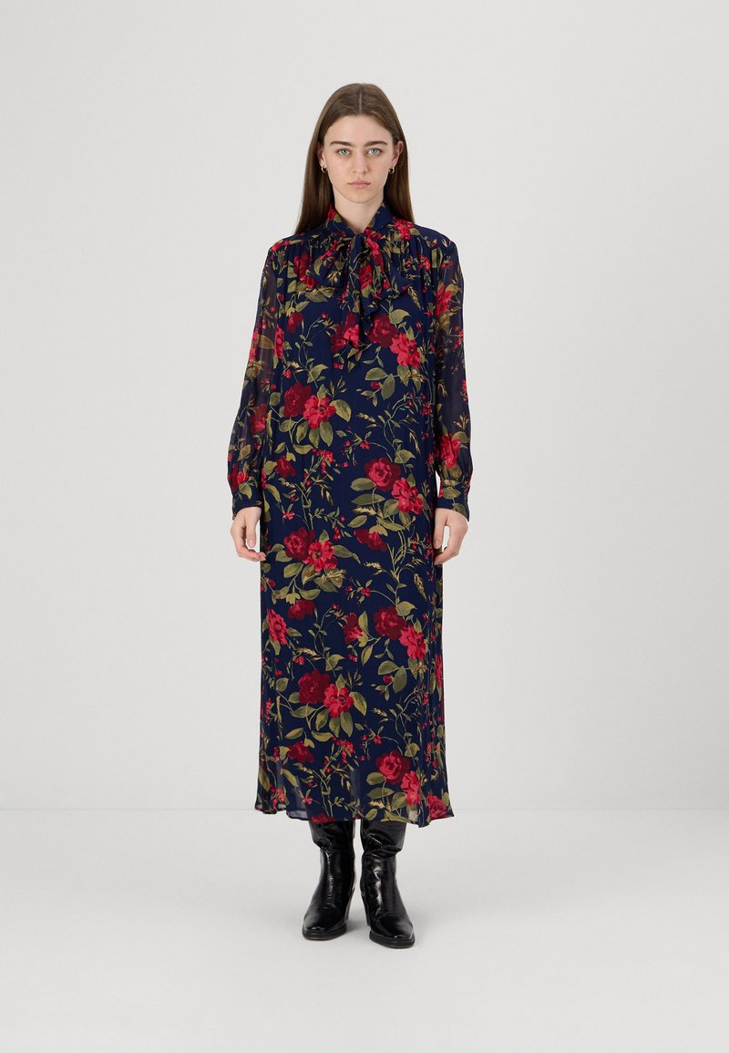 Long floral dress in navy with red and green patterns. Features a neck tie and sheer sleeves, complemented by black ankle boots.