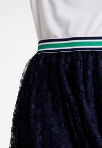 Granatowa spódnica z tiulu w wzory, zestawiona z dopasowanym białym topem, z paskiem elastycznym w paski w kolorze granatowym, zielonym i białym.