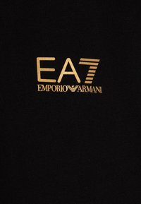 Crni materijal s zlatnim logotipom "EA7" i "EMPORIO ARMANI." Dizajn uključuje horizontalne pruge i glatku teksturu.