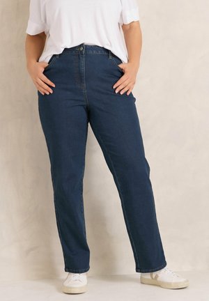 Person trägt dunkelblaue Jeans mit hoher Taille, die Hände in den Taschen, kombiniert mit weißen Turnschuhen und einem weißen kurzärmligen Hemd.