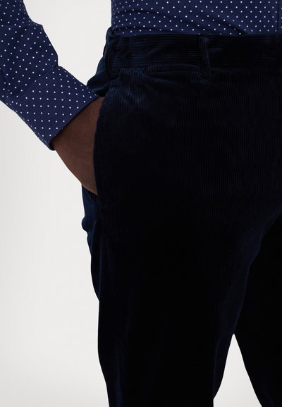 Marineblå kordfløyelsbukser med teksturert overflate, kombinert med en marineblå skjorte med prikkemønster. Har standard linning og sidelommer.