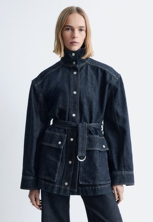 Donkere spijkerjack met een hoge kraag, drukknoopsluiting en een tailleband met ceintuur. Heeft een ontspannen pasvorm en twee grote voorzakken.