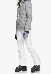 Graue isolierte Jacke mit Kapuze, weiße Skihose, gemusterte blaue Handschuhe und schwarze Skischuhe. Verfügt über ein gestepptes Design und Reißverschlüsse.