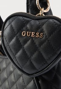 Borsa a tracolla nera a forma di cuore con motivo trapuntato e logo "GUESS" dorato. Include dettagli in metallo dorato e cerniera.