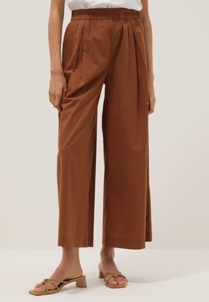 Mujer con pantalones marrones de talle alto y pierna ancha con pliegues, combinados con sandalias beige de tacón bajo y una camiseta blanca metida dentro.