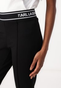 Črne hlače z gladko teksturo imajo širok pas, označen z "KARL LAGERFELD" in belimi črtami, prikazane na beli podlagi.