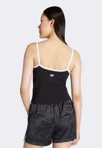 Černý top bez rukávů s bílými ramínky, který obsahuje malé bílé logo. V kombinaci s černými šortkami s proužky, které mají uvolněný střih a strukturu.