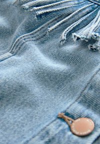 Zbliżenie jasnoniebieskiego denimu z widocznymi przeszyciami, postrzępionymi krawędziami oraz małym, okrągłym, brązowym guzikiem przymocowanym sznurkiem.