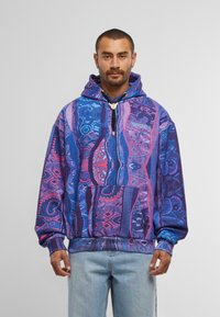 Uomo che indossa un hoodie a motivi vivaci blu e viola con tasca frontale e jeans blu chiaro, in piedi contro uno sfondo grigio semplice.