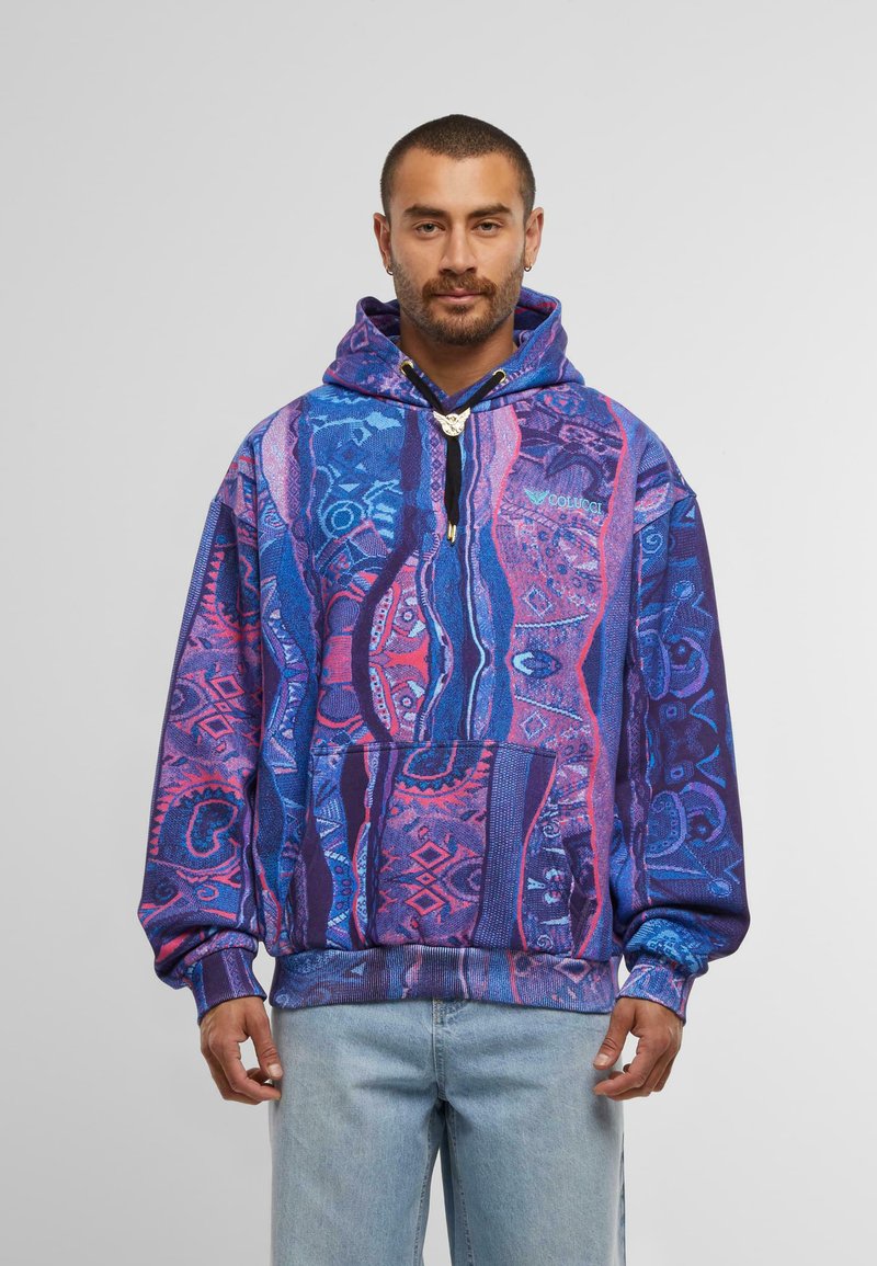 Uomo che indossa un hoodie a motivi vivaci blu e viola con tasca frontale e jeans blu chiaro, in piedi contro uno sfondo grigio semplice.